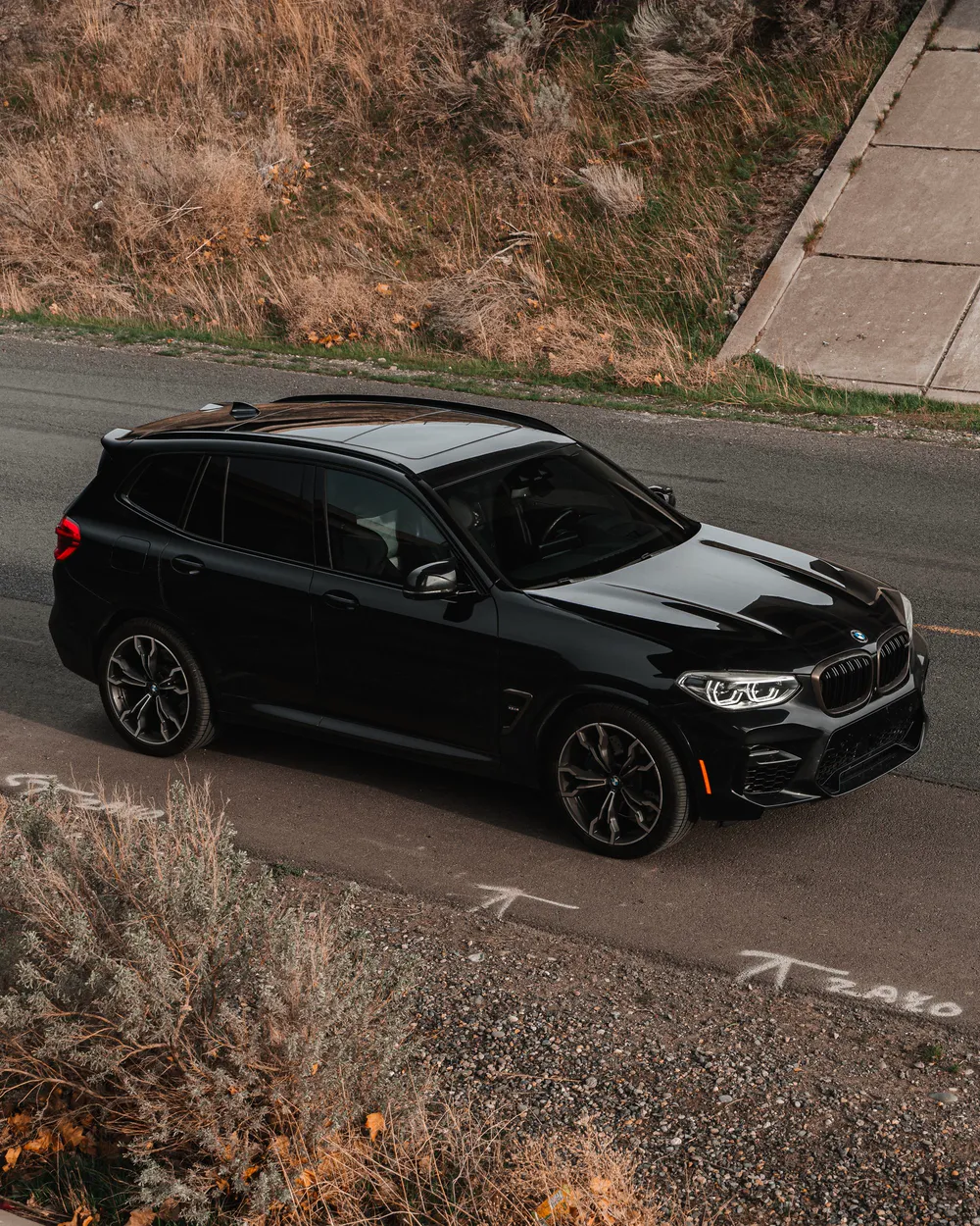 A black BMW X3 at the edge of a road, seen from above beside dry grass