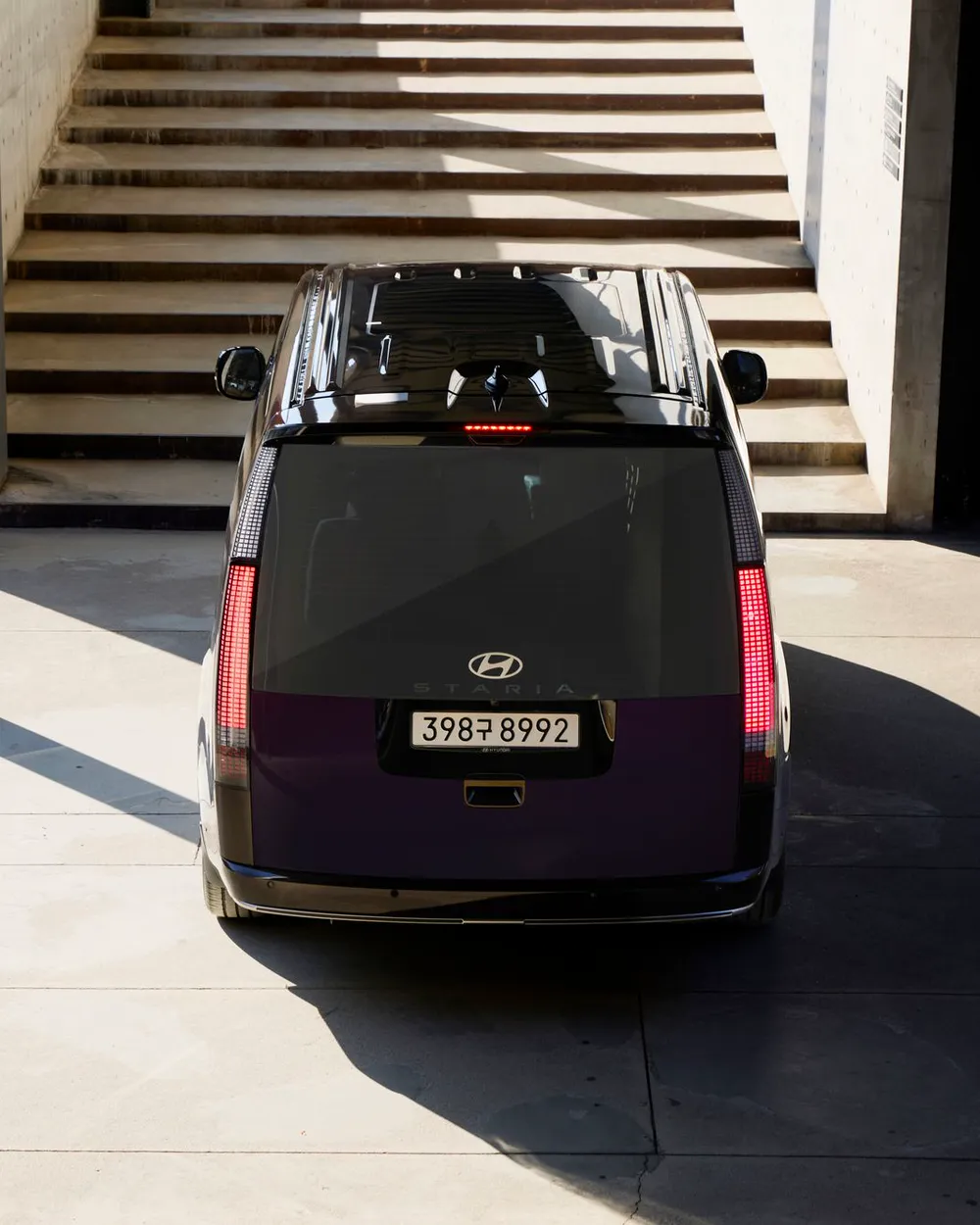 A dark Hyundai Staria seen from behind, parked at the foot of a sunlit concrete stairway